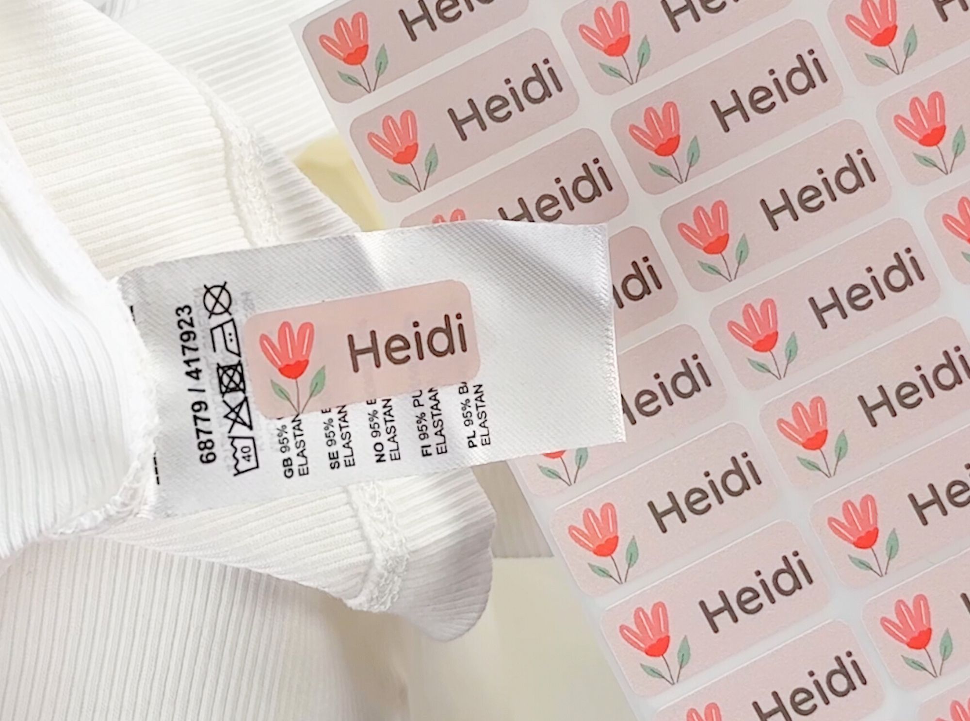 Namensaufkleber und bügeletiketten – personalisierte lösungen für jeden bedarf 3 Aufkleber mit dem namen "heidi" und einer roten blume. Im hintergrund ein set mit den namensaufklebern.