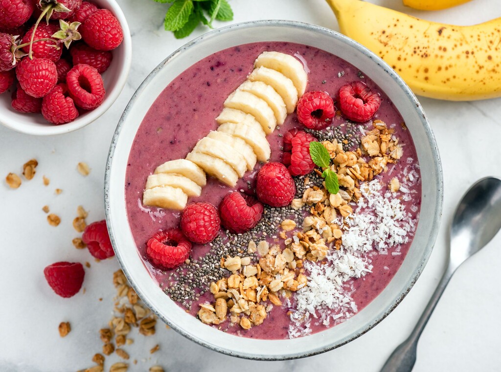 Smoothie-bowl mit frischen himbeeren, bananenscheiben, chiasamen, granola und kokosraspeln, gesunde frühstücksidee