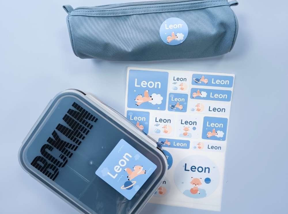 Blaue Namensaufkleber mit einem kleinen Fuchs im Weltraum und dem Text "Leon" auf verschiedenen Schulutensilien.
