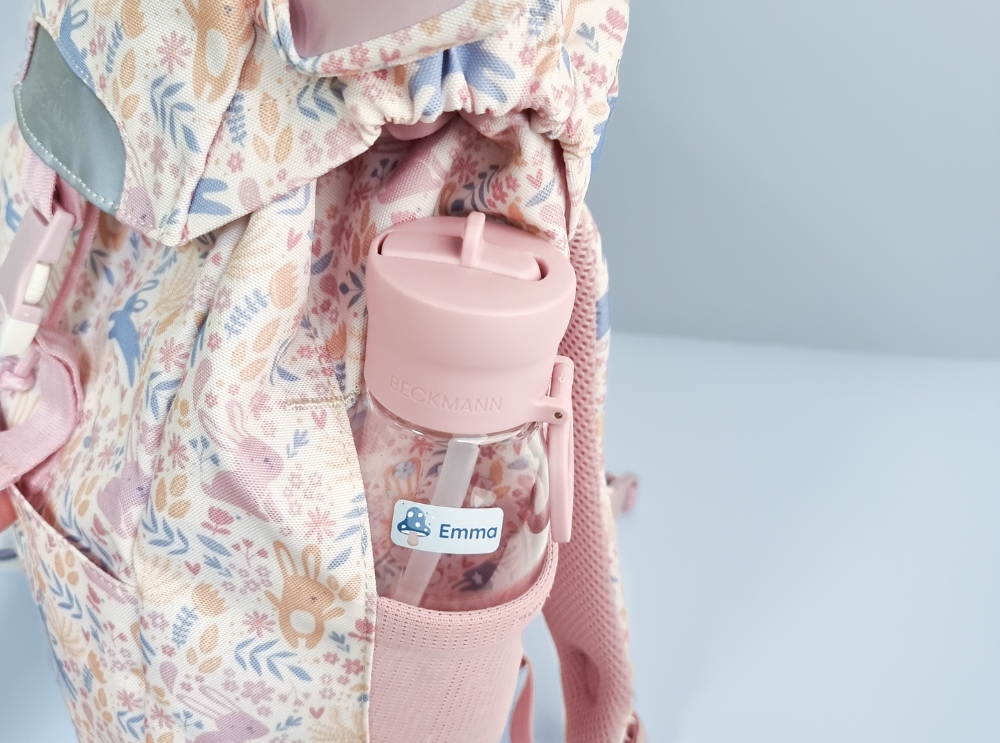 Nahaufnahme einer rosa trinkflasche mit namensaufkleber in der seitentasche eines geblümten rucksacks